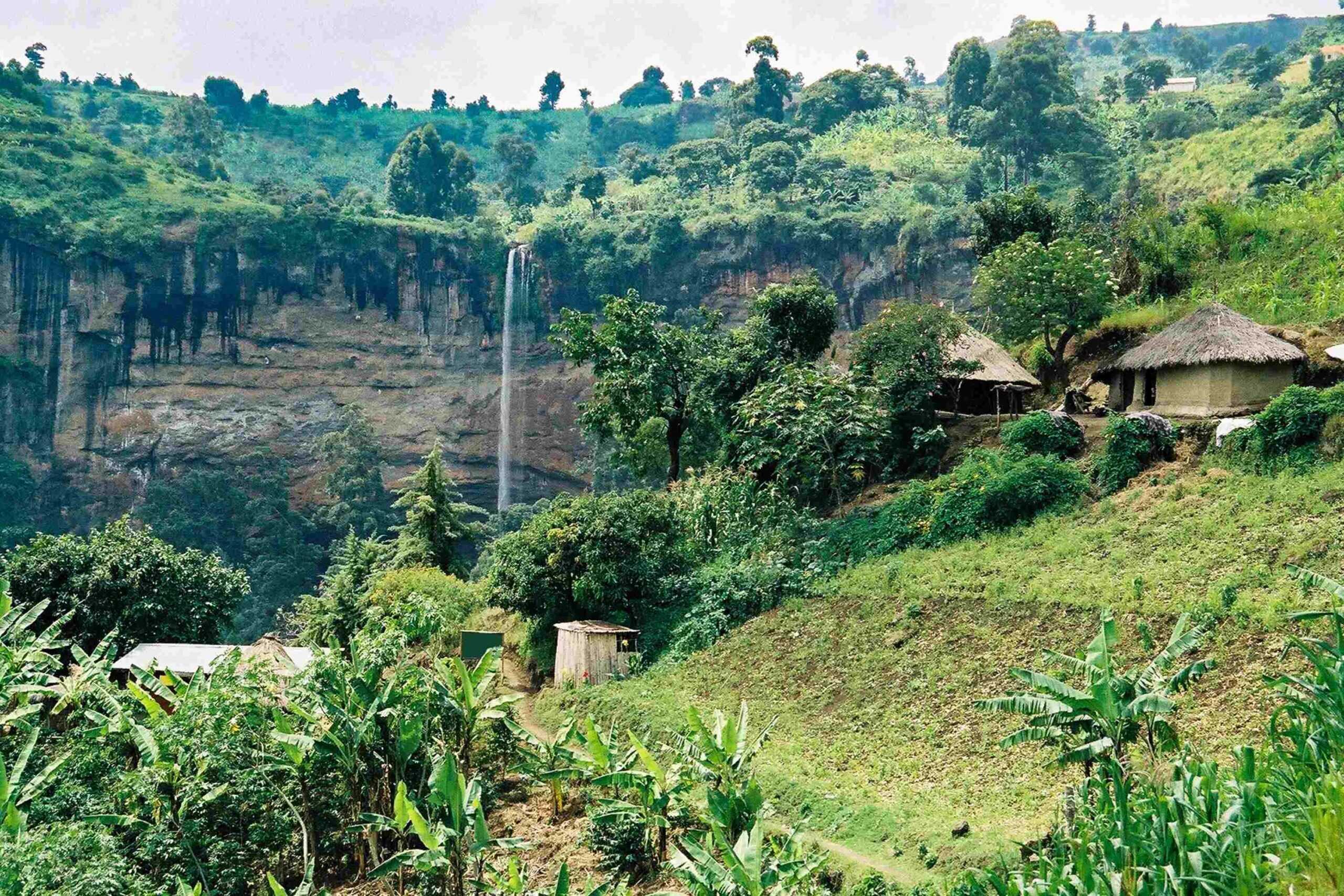 Grüne, hügelige Landschaft mit Hütten und einem hohen Wasserfall im Hintergrund