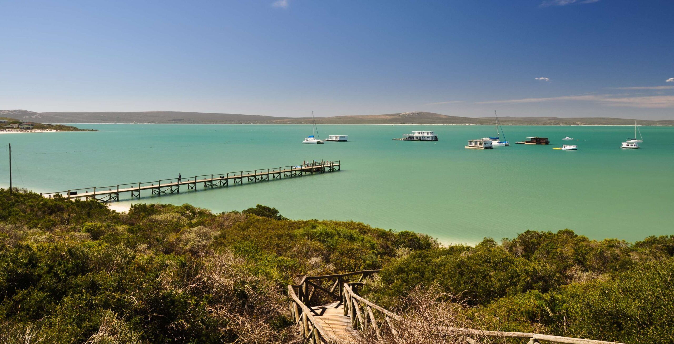 Treppen führen hinunter zur Langebaan-Lagoon mit Holzsteg im türkisfarbenen Wasser und Schiffen