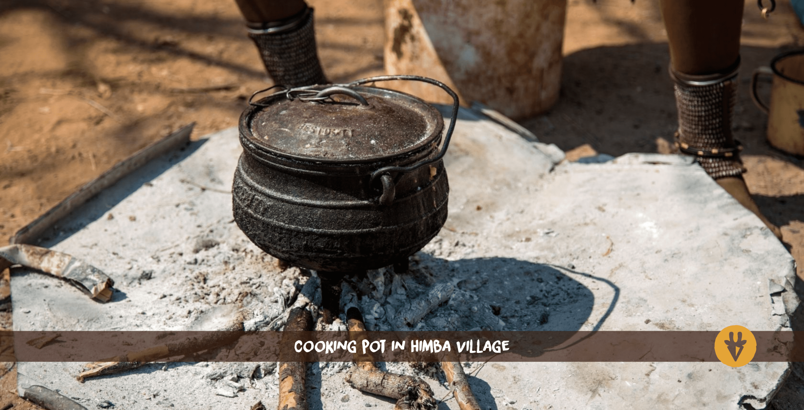 Ein dunkelbrauner Topf mit einem Deckel und Henkel steht auf Asche und kleinen Ästen eines erloschenen Feuers in einem Himba-Dorf